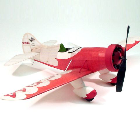 Dumas Gummimotorflugzeug im Retro-Design mit rotem Propeller und bunten Details, präsentiert aus seitlicher Perspektive.